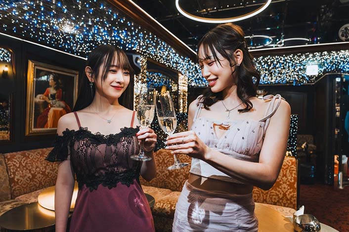 Two young women dressed elegantly, smiling and clinking champagne glasses in a festive, dimly lit lounge with decorative string lights.