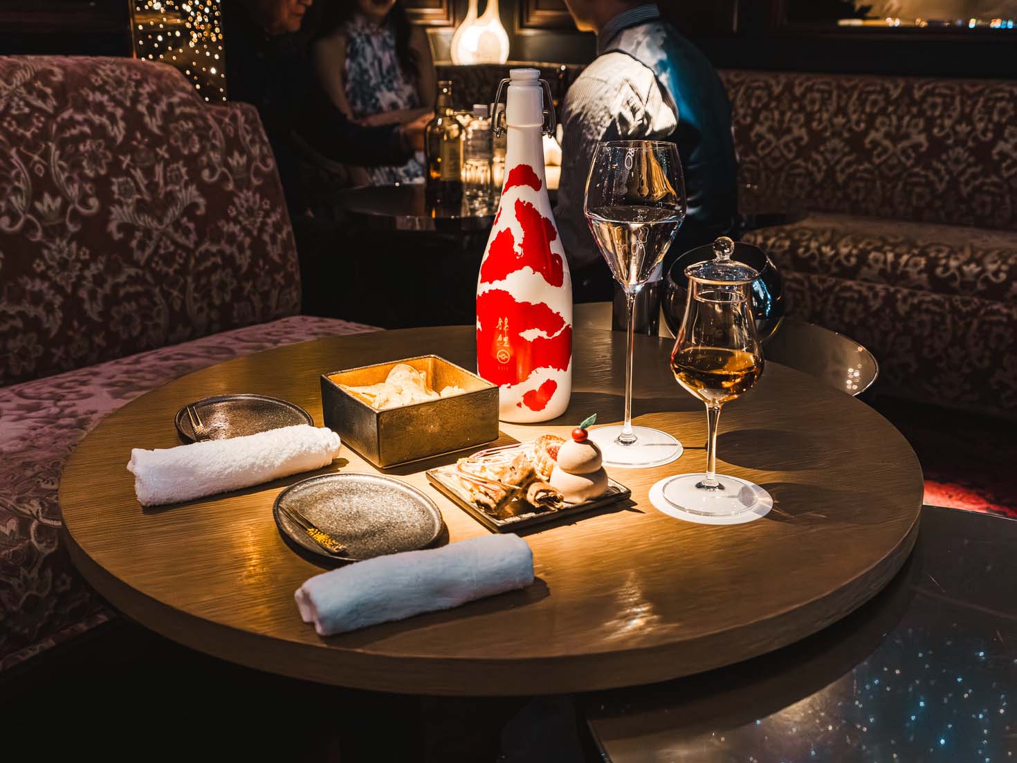 Round wooden table set with two rolled white napkins, two empty plates, a white bottle with red design, two glasses with drinks, and a wooden tray with assorted desserts in a dimly lit lounge.