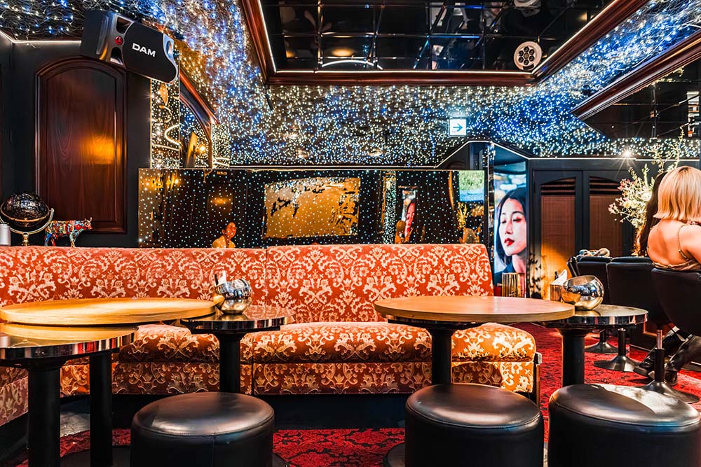 Luxurious lounge with patterned orange sofa, round wooden tables, black stools, and twinkling string lights on mirrored walls and ceiling.