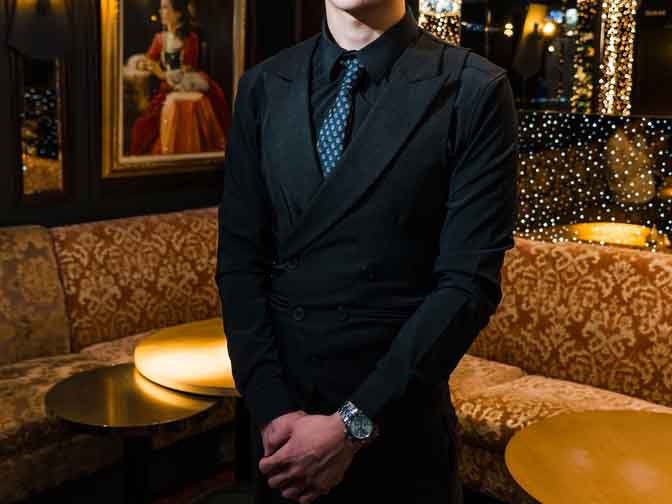 Man in a black suit and patterned blue tie standing in a dimly lit lounge with patterned brown couches and framed artwork on the wall.